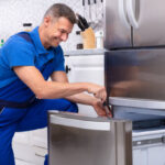Mature Male Serviceman Repairing Refrigerator With Toolbox In  Kitchen